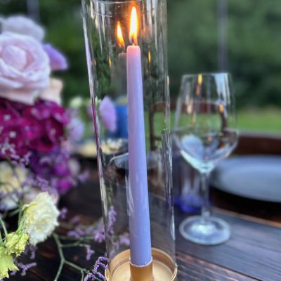 Detailaufnahme eines Tischarrangements mit lilafarbenen Gläsern, Kerzenzylindern und sommerlicher Floristik in Gelb und Violett.